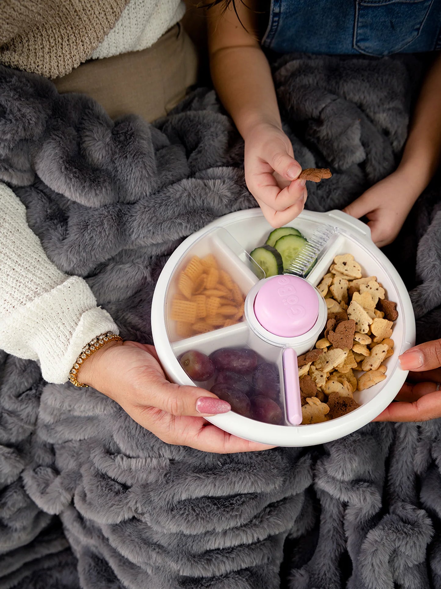 GoBe Snack Spinner in lilac with assorted snacks, held by hands under a cozy blanket.